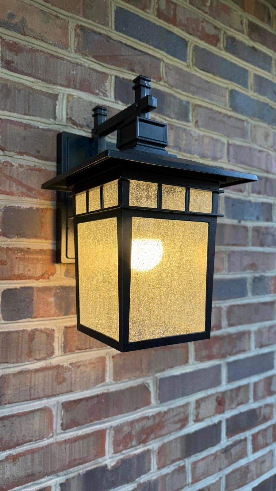 Craftsman style wall sconce lit on a brick exterior wall